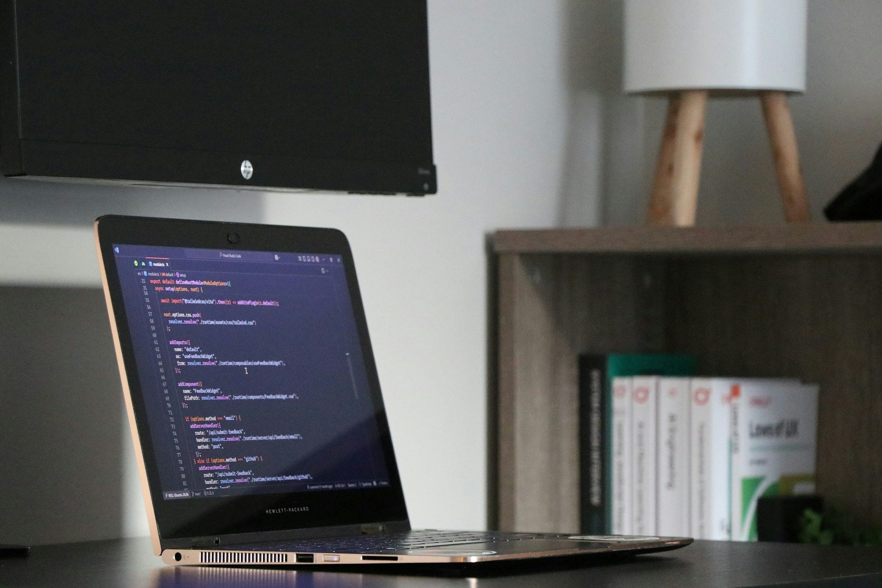 A laptop with code open beside books and a desk lamp